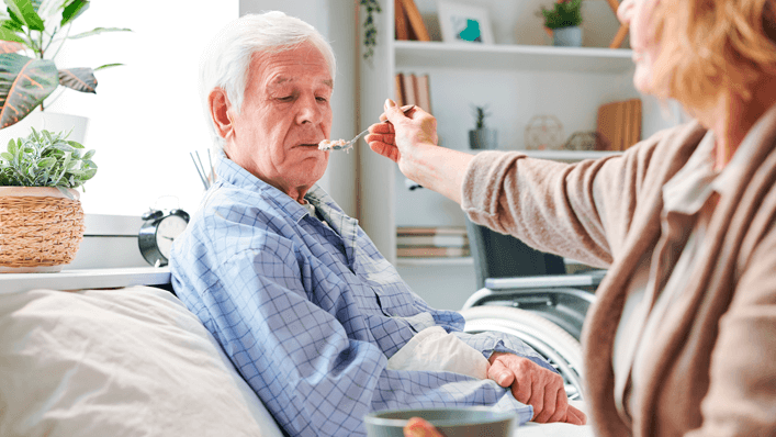 Mulher alimentando idoso de cama - colesterol